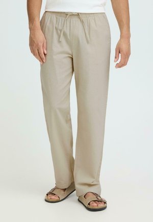 Man wearing beige drawstring pants and beige sandals standing against plain background.