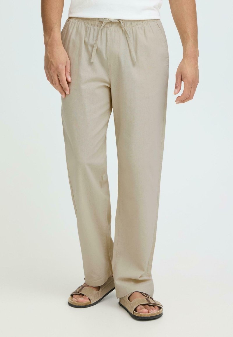 Homme portant un pantalon beige à cordon de serrage et des sandales beiges, debout devant un fond uni.