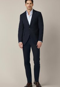 Marynarka w kolorze granatowym, o dopasowanym kroju, z klapami typu notch i dwoma guzikami. W komplecie wąskie spodnie i jasnoniebieska koszula. Brązowe buty.