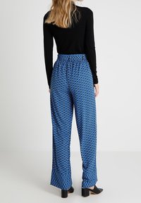 Blå mönstrade byxor med vida ben och elastisk midja; bakvy visar flödande tyg och subtila vita diamantformer. Svart långärmad topp.