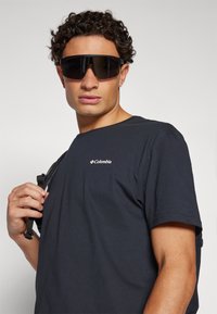 Tricou din bumbac de culoare albastru marin cu un mic logo alb Columbia. Modelul poartă ochelari de soare supra-dimensionați negri și ține o curea neagră pe umăr.