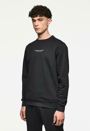 Man met krullend haar, gekleed in een zwart sweatshirt en bijpassende broek met de tekst "WEEKEND OFFENDER ADMIT NOTHING" op de borst en het been.