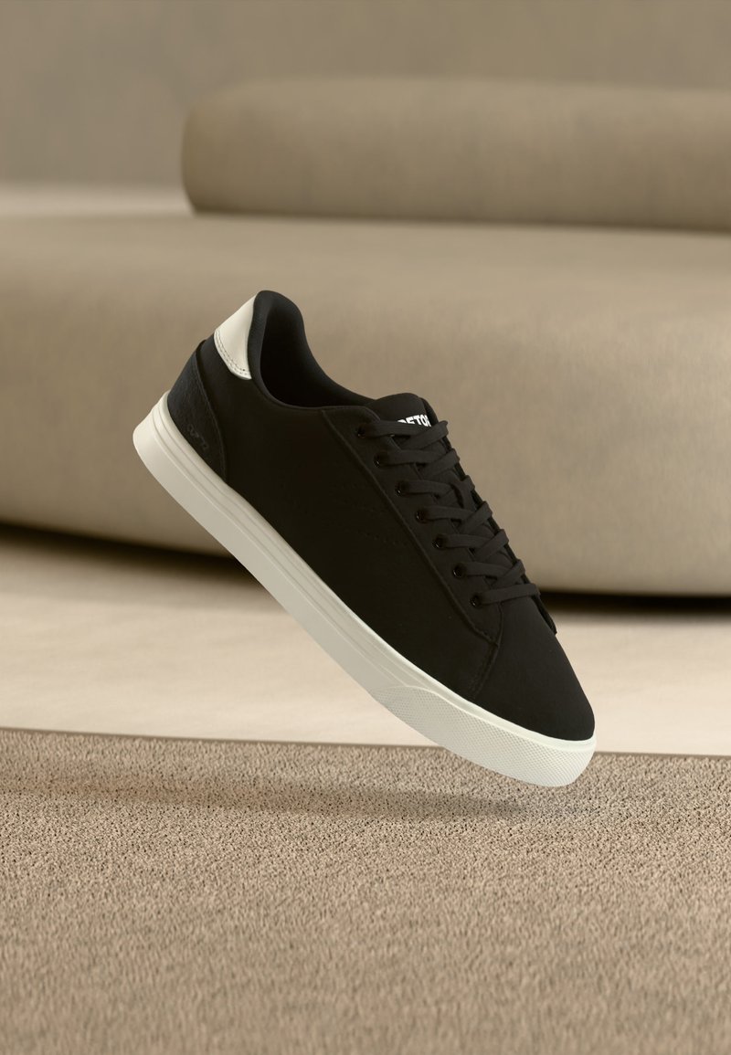 Μαύρο αθλητικό παπούτσι με λευκή σόλα και λεπτομέρεια στη φτέρνα, που αιωρείται πάνω από μπεζ χαλί με καθιστικό σε ουδέτερο τόνο θολωμένο στο φόντο.