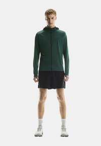 Junger Mann steht, trägt einen dunkelgrünen Zip-Hoodie, schwarze Shorts, weiße Socken und weiße Laufschuhe vor einem schlichten Hintergrund.
