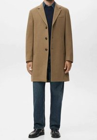 Manteau en laine marron à coupe ajustée, avec trois boutons, poches avant, porté sur un pull marine et un jean bleu, associé à des chaussures noires.