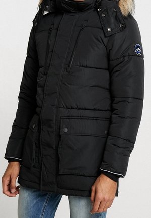 Doudoune noire avec une capuche bordée de fourrure, poches zippées, fermeture à boutons-pression et poignets côtelés. Présente un patch logo sur le bras.