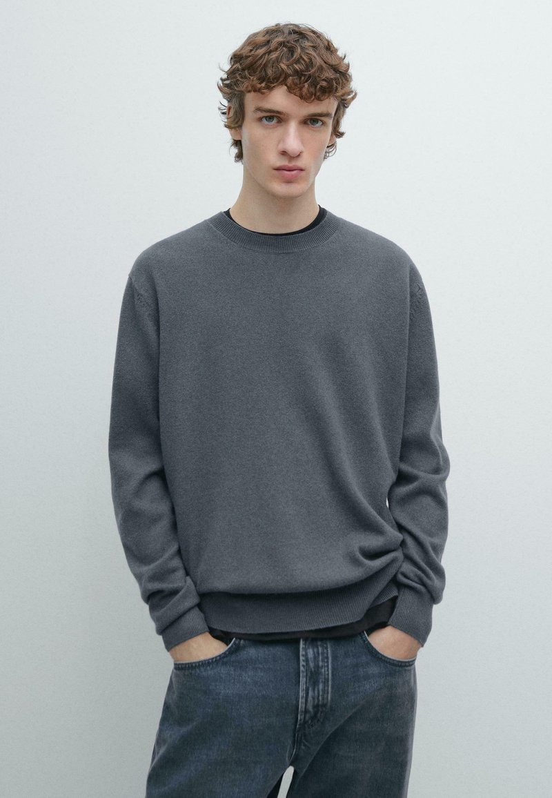 Gray crewneck sweater in soft fabric, ribbed cuffs and hem, worn over a black shirt, paired with dark denim jeans. Simple, clean design.
