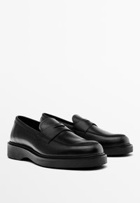 Schwarze Lederloafers mit glatter Oberfläche, abgerundeter Spitze und einer dicken, schwarzen Sohle. Verfügt über einen dekorativen Riemen über dem Obermaterial.