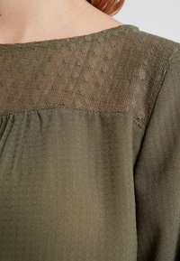 Top in tessuto verde con texture e pannello trasparente decorato all'escella, caratterizzato da punti sottili e un design morbido e fluido.