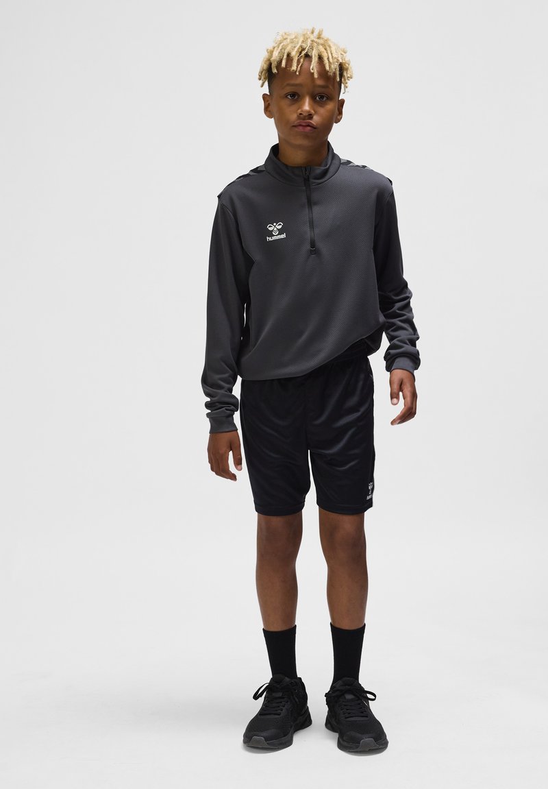 Jongen met lichtblonde dreadlocks die een zwart sportshirt met lange mouwen, zwarte shorts, zwarte sokken en zwarte sneakers draagt, staand tegen een effen achtergrond.