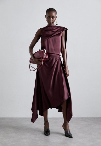 Maroon satin dress with draped detailing, asymmetrical hemline. Model holds a matching bag. Black pointed heels with clear block heels.