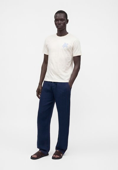Homme debout, une main dans la poche, portant un t-shirt crème avec un motif de tortue, pantalon bleu marine et sandales marron foncé.