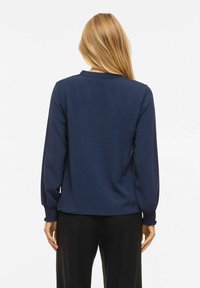 Femme aux longs cheveux blonds portant un chemisier texturé bleu marine à manches longues et un pantalon noir, dos tourné à la caméra sur un fond blanc.