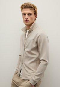 Chaqueta cremosa de zip con cuello alto, ajuste slim y puños acanalados. Textura suave con un diseño minimalista y detalle de costura frontal.
