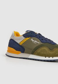 Zapatilla en azul marino y verde olivo con texturas de ante y malla, con acentos naranjas y suela blanca. Detalle del logo en el lateral.