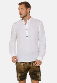 Weißes Leinenhemd mit langen Ärmeln und Knopf-Henley-Ausschnitt, kombiniert mit braunen Lederhosen, die dekorative Akzente und Stickereien aufweisen.