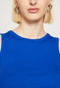 Top de tirantes azul con textura acanalada y escote redondo. La modelo lleva pendientes de aro plateados y tiene el cabello rubio corto. Textura de tela suave.