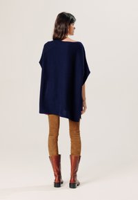 Navyblauw gebreid poncho met een relaxte pasvorm, zijsplitsingen en een gladde textuur, gedragen over bruine suede leggings en bruin kniehoge laarzen.