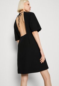 Μαύρο φόρεμα με ανοιχτή πλάτη, κοντά μανίκια και ένα σφιχτό αξεσουάρ στον λαιμό. Φτιαγμένο από υφασμάτινη υφή με χαλαρή εφαρμογή.