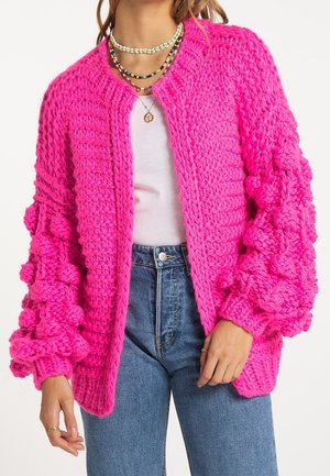 Femme portant un cardigan tricoté épais rose vif avec des manches bouffantes texturées, un haut blanc, un jean bleu et des colliers de perles superposés.