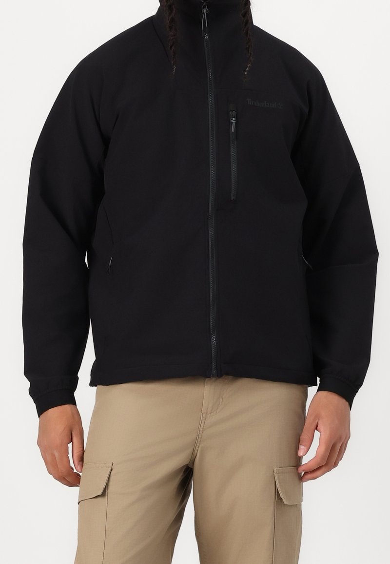 Homme portant une veste Timberland noire zippée et un pantalon cargo beige sur un fond uni.