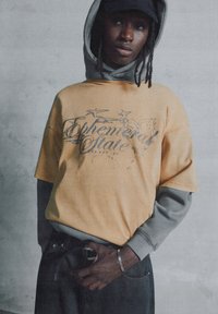 Gul grafisk t-shirt med "Ephemeral State" tryk, lagd ovenpå en grå hættetrøje. Mørke jeans, tætsiddende med et bælte, og et håndledsarmbånd.