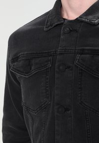 Záběr zblízka na osobu nosící černou džínovou bundu s zapnutými kapsami na knoflíky na hrudi a viditelnými šicími detaily.