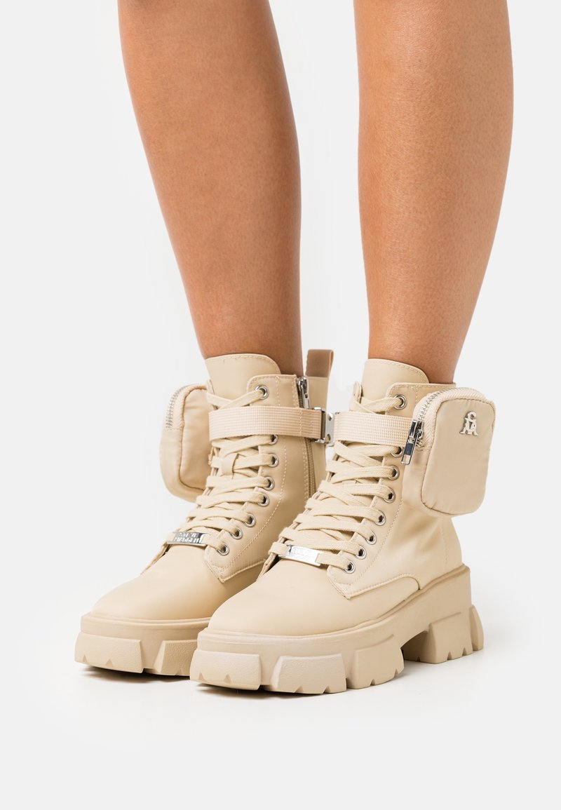 Bottes de combat à plateforme épaisses beiges avec poches à fermeture éclair latérales, portées sur des jambes nues sur un fond uni.