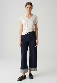 T-shirt a scollo a V crema abbinato a jeans a gamba larga blu scuro con risvolto in denim. Il modello indossa mocassini marroni scuri.