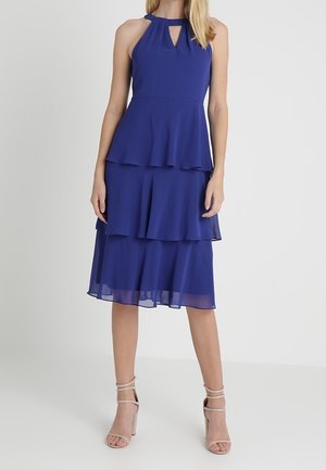 Kobieta w bezrękawnikowej, królewskiej niebieskiej sukience midi z warstwami i dekoltem z wycięciem oraz w cielistych sandałach na wysokim obcasie z paskami, stojąca na białym tle.