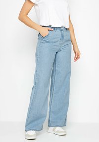 Jeans de mezclilla de pierna ancha en azul claro con bolsillos frontales y cierre de botón. Combinados con una camiseta blanca y zapatillas blancas.