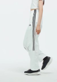 Biele športové nohavice s čiernym trojpruhovým designom pozdĺž strán, s elastickým pásom a voľným strihom, skombinované s čiernymi teniskami.