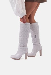 Stivali bianchi al ginocchio con design perforato, dettaglio con fibbia e punta a forma di coltello su un tacco in legno. Texture liscia e silhouette slanciata.