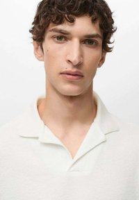 Polo bianco con tessuto testurizzato, caratterizzato da un colletto pieghevole e una patta senza bottoni. Il modello ha i capelli corti e ricci e un'espressione neutra.