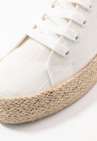 Sneaker bianca in tela con suola piatta in juta spessa. Presenta lacci bianchi piatti e occhielli in metallo. Superficie testurizzata sulla scarpa.