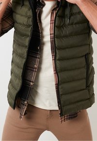 Olivově zelená prošívaná vesta s prošívanou texturou, nošená přes kostkovanou košili s knoflíky. Béžové tričko pod ním a na pohled světlehnědé slim-fit kalhoty.