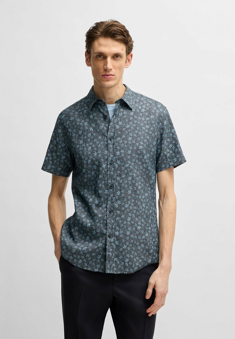 Camicia a maniche corte con bottoni in tessuto blu scuro con un motivo floreale blu chiaro. Presenta un colletto alla francese e sette bottoni lungo la parte anteriore.