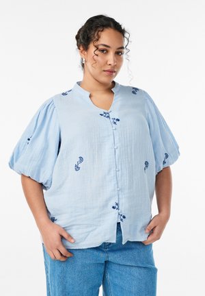Kobieta z kręconymi włosami, ubrana w jasnoniebieską bluzkę z bufkami i kwiatowym haftem, w połączeniu z niebieskimi dżinsami, stojąca z rękami w kieszeniach.