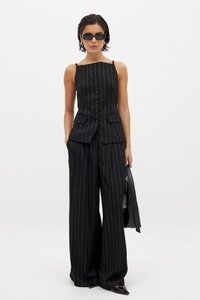 Črn prugast krojen brezrokavnik z gumbi, v kombinaciji s širokimi črnimi hlačami. Uporabljen z elegantnimi črnimi sončnimi očali in v roki drži torbico.