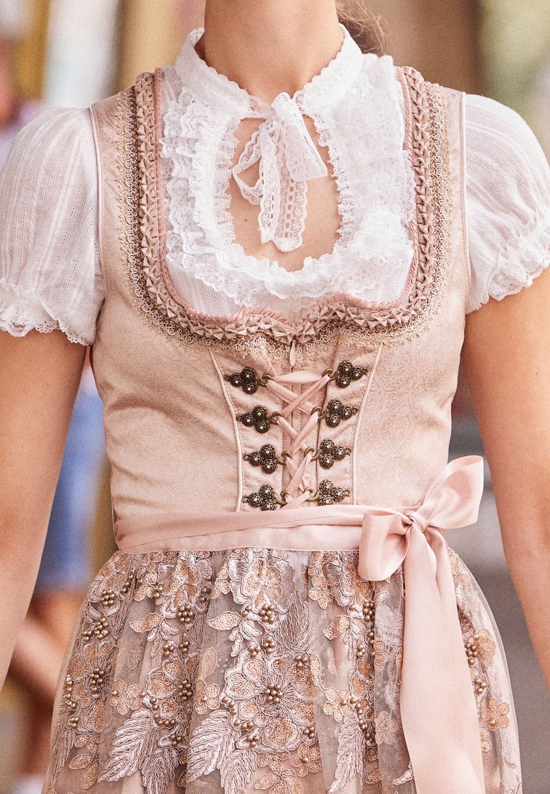 Femme portant une robe traditionnelle dirndl rose avec un corsage à lacets, un chemisier blanc à manches bouffantes, un col en dentelle et un tablier brodé noué avec un nœud en satin.