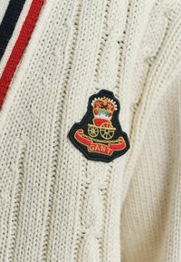 Cremfarbener Kabelstrickpullover mit rot-, weiß- und blauen Streifen sowie einem schwarzen Patch mit einer Krone, einer Kutsche und dem in Gold und Rot gestickten "GANT"-Text.