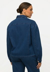 Jeansjacka i marinblått tyg, med vertikala vita ränder och en strukturerad krage. Har knäppta manschetter och kontrastsömmar.