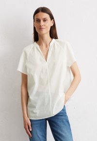 Weiße Bluse mit kurzen Ärmeln und V-Ausschnitt, aus leichtem Stoff gefertigt. Sie hat eine lockere Passform und dezente Nähte. Kombiniert mit blue Jeans.