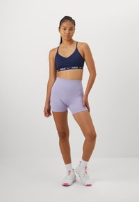 Musta urheilu-toppi, jossa on valkoinen Nike-logo alareunassa, yhdistettynä vaalean lilaan korkeavyötäröiseen shortsipariin. Mallilla on päällään valkoiset tennarit, joissa on vaaleanpunaisia yksityiskohtia.