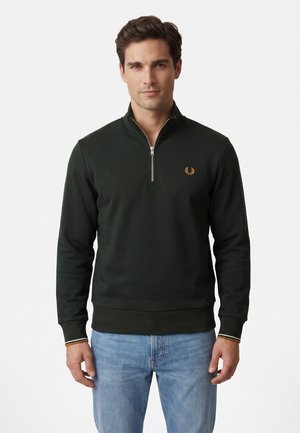 Hombre que lleva un suéter verde oscuro con cremallera hasta el pecho y un pequeño logo naranja en el pecho, y vaqueros azul claro, de pie frente a un fondo liso.