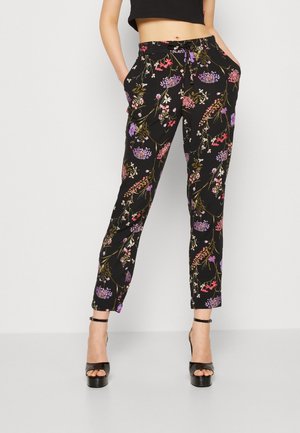 Femme portant un pantalon slim noir à fleurs, un top court noir et des sandales à talons hauts noires, debout sur un fond blanc.