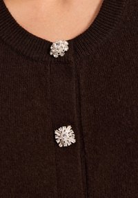 Ciemnobrązowy kardigan z prążkowanym dekoltem, z dwoma ozdobami: jedną w kształcie okręgu i drugą w kształcie gwiazdy, obie w srebrnych odcieniach.