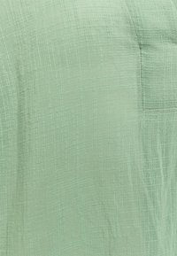 Tessuto verde chiaro, strutturato, con un motivo a griglia. Include un piccolo dettaglio a tasca, che sottolinea un design rilassato e traspirante.