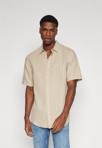 Chemise beige à manches courtes et boutonnières, fabriquée en tissu léger, avec une poche sur la poitrine gauche, portée avec un jean bleu.