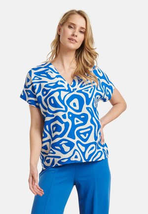 Short-sleeve blouse in a light fabric, featuring a bold blue abstract pattern on a white background. V-neck design with a loose fit.